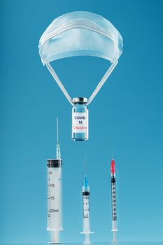 The vaccine labeled COVID-19 is dropped on a scraper from a protective medical mask, on a blue background with syringes. Isolate. The concept of saving the planet's population from viruses and infections. Thrown off the plane.