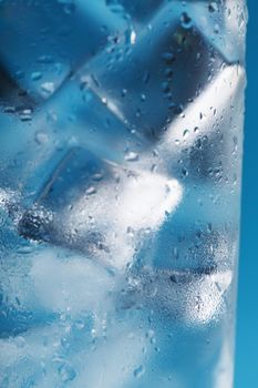 Ice cubes in a misted glass with drops of ice water close-up macro. Refreshing and chilling drink in hot weather. Blue background. Soft Selective Focus