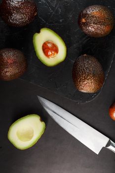 Whole and sliced avocados with a knife on a black textured black table, flat masonry. Top view
