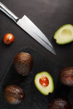 Whole and sliced avocados with a knife on a black textured black table, flat masonry. Top view