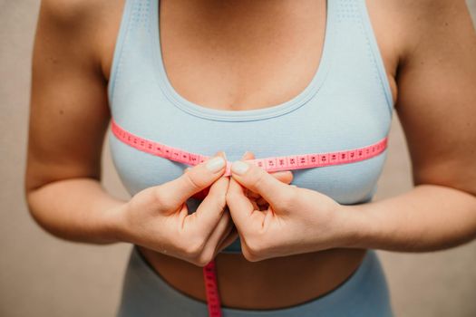 Cropped view of slim woman measuring breasts with tape measure at home, close up. Unrecognizable European woman checks the result of a weight loss diet or liposuction indoors. Healthy lifestyle..