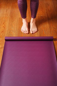 The girl's feet stand in front of an unfolded Yoga mat on the wooden floor. Yoga practice classes, asana exercises