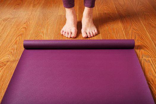 The girl's feet stand in front of an unfolded Yoga mat on the wooden floor. Yoga practice classes, asana exercises