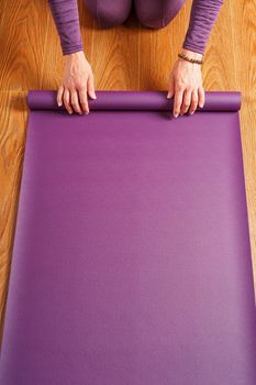 A woman lays out a lilac yoga mat on the wooden floor. Women practicing yoga. A healthy lifestyle in isolation.