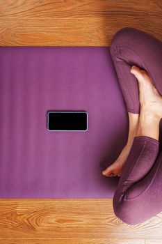 A girl does yoga with a smartphone online via the Internet on a lilac rug in the living room.