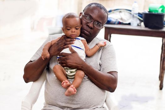 a young man sitting in a white chair in glasses and gray t-shirt with his baby on his chest looking at the camera.