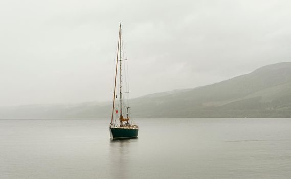 Landscape view of Loch Ness with calm water, mist and hills in the background and a sailboat with sails up floating peacefully on the water.