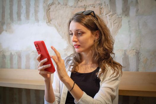 Young woman in white shirt and black top is browsing using her red smartphone to make online shopping or payment sitting indoors on loft wall background, technology, happy people concept.