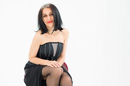 Middle aged woman starting getting grey-haired is posing in studio while sitting on the chair in black dress and stockings on white background, middle age skincare cosmetics, cosmetology concept.