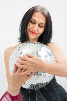 Studio photo of middle aged woman starting getting grey-haired wearing black clothes with silver disco ball in hands on white background, middle age sexy lady, happy life concept.