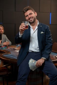 Cheerful stylish young bearded man leaning on gaming table in casino, holding fan of banknotes and raising glass of whisky for successful game. Gambling concept