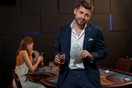 Slightly drunk young bearded man winning in poker, standing near gaming table in casino with glass of brandy and fan of banknotes. Leisure, lifestyle, gambling concept