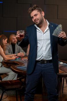 Successful bearded guy breaking bank in poker game, standing with banknotes and glass of alcohol near gaming table with opponents. Good luck gambling concept