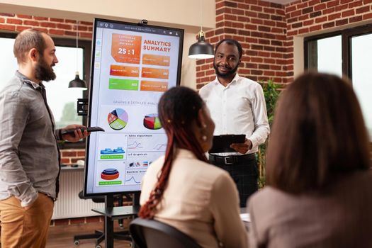 African american entrepreneur showing business graph discussing management statistics during financial meeting in startup office. Diverse businesspeople planning company partnership