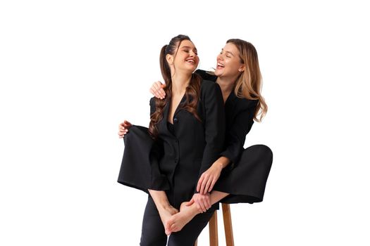 Two young business woman dressed black suit laugh and happy. One woman sitting chair and hugging other woman with barefoot legs. Positive caucasian female business people look at each other. Isolated