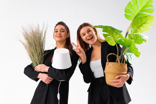 Two business woman dressed black suit smile holding houseplant and lamp standing isolated on white background. Moving concept. Relocation new office with positive. One woman shoving victory sign
