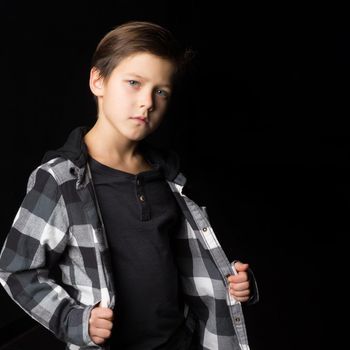 Close up portrait of boy wearing casual clothes. Handsome preteen boy in plaid black and gray shirt standing against black background looking seriously at camera