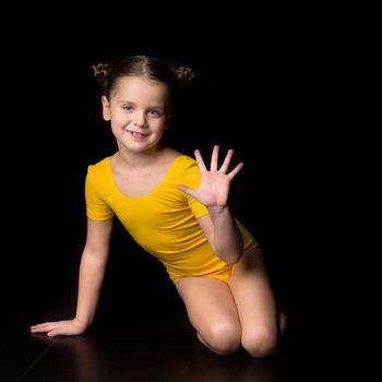 Cute cheerful little girl gymnast sitting on floor. Smiling kid wearing yellow leotard sitting on floor showing five fingers against isolated black background in studio