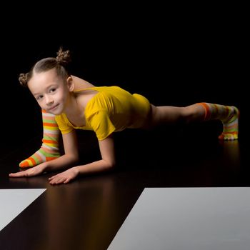 Cute flexible little girl doing elbow plank exercise. Girl acrobat wearing yellow leotard and striped socks doing gymnastics exercise against isolated black background in studio