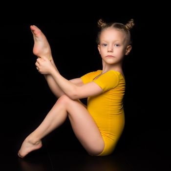 Girl sitting on the floor raising her leg up. Sportive barefoot kid gymnast or acrobat wearing yellow leotard performing acrobatic exercise against isolated black background in studio