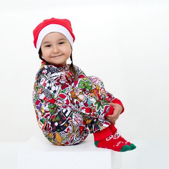 Happy girl in Christmas costume sitting on floor. Cute little girl wearing colorful overalls, red socks and Santa Claus cap sitting on floor hugging her knees against isolated white background