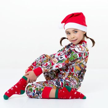 Cute girl with pigtails in overalls and Santa cap. Cheerful little girl wearing colorful Christmas costume sitting on floor sideways to camera on white background