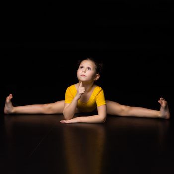 Cute young girl sitting on cross or transverse splits. Beautiful barefoot kid wearing yellow leotard performing rhythmic gymnastic exercise and looking up thoughtfully against black background
