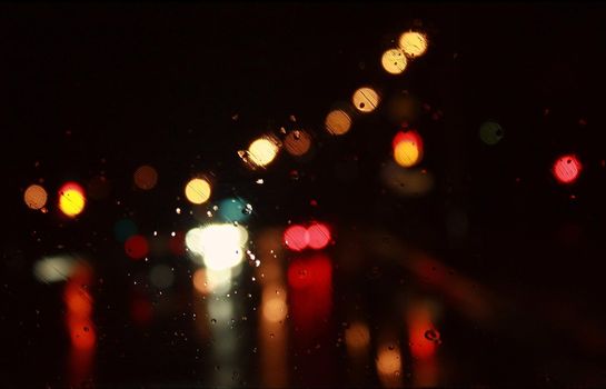 Car traffic lights bokeh through rainy window glass on black background. Defocused city night colorful round lights. 