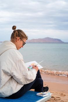 Hobbbies and leisure. young woman artist painting or doing travel sketches using watercolor sitting on the beach