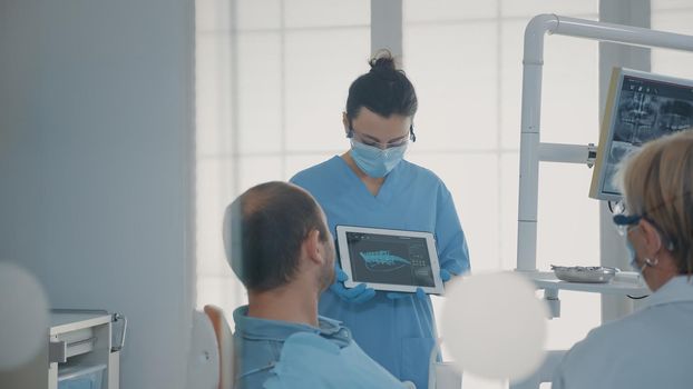 Orthodontic nurse holding x ray results on tablet to show and explain diagnosis to patient in dentistry office. Team of experts showing teeth radiography on device, talking about implant.