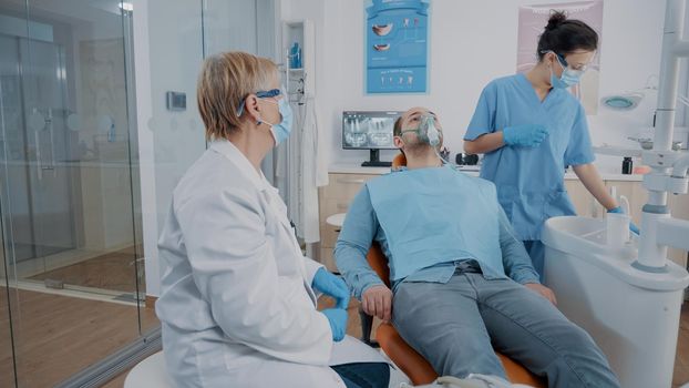 Dentistry assistant putting oxygen mask on face of patient to do anesthesia before surgical procedure. Oral care specialist giving anesthesic to man with toothache and using drill tool.