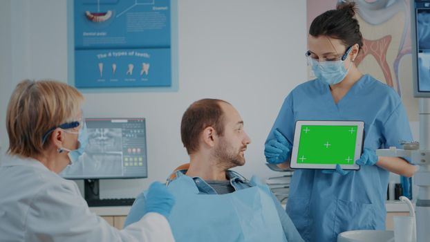 Patient and dentistry team looking at tablet with green screen. People in stomatology cabinet using isolated template with mockup copy space and chroma key background on digital device.