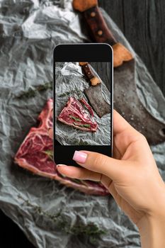 Photographing food concept - woman takes picture of raw dry aged t-bone steaks for grill with fresh herbs and cleaver. Top view