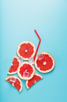 A composition of grapefruit slices and ice cubes with a straw on a blue background in the form of a refreshing drink. Top view, free space