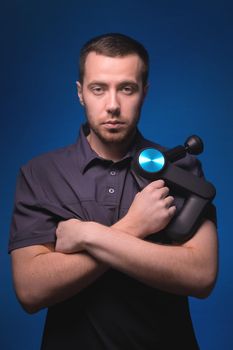 Portrait of a confident professional masseur with a percussion massager in his hands. Low key, blue background. Shock wave massage.