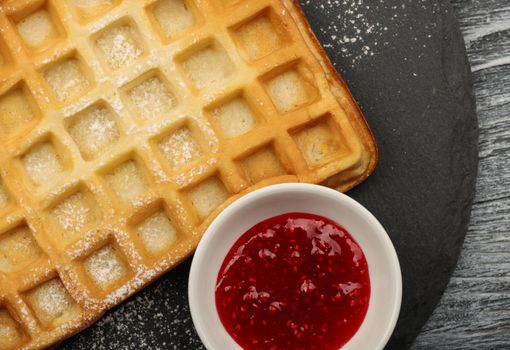 Close up big sweet traditional American or Belgian Brussels waffle with powder sugar and raspberry jam served on black slate board on table, elevated top view, directly above