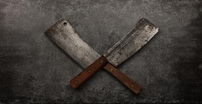 Close up two crossed vintage butcher meat cleavers on cutting board or grunge gray stone table surface with copy space, elevated top view, directly above