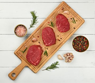 Close up three aged prime marbled raw tenderloin or fillet mignon beef steaks on brown oak wood cutting board, with spices, over white wooden table background, elevated top view, directly above