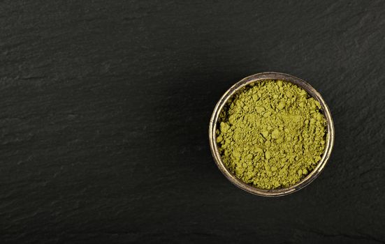 Close up metal bowl full of ground unroasted raw green Arabica coffee on black slate board background, elevated top view, directly above