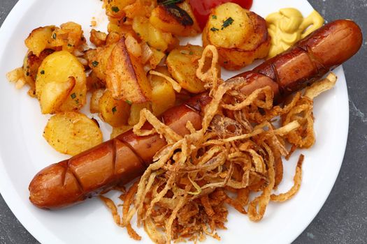 Close up portion of one big grilled sausage with homemade roasted potato, fried onion rings, ketchup and mustard on white plate, elevated top view, directly above