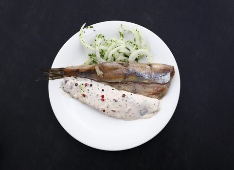 Close up portion of salted pickled herring fish filets with onions and sauce on white plate over black table, elevated top view, directly above