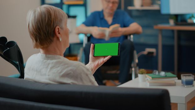 Mature woman holding horizontal green screen on smartphone, having physical disability. Senior person looking at isolated copy space template and chroma key mock up background on display.