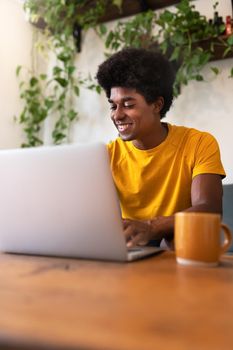Happy young African American man sitting on sofa using laptop in the morning to check his social media. Vertical image. Technology concept. At home concept.