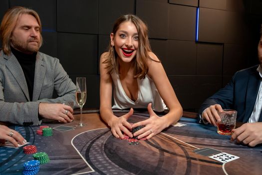 Happy excited young brown-haired woman sitting at poker table in casino, raking pile of won chips satisfied with successful game. Concept of passion for gambling