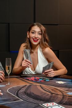 Cheerful lucky brown-haired girl winning in poker, sitting at gaming table in casino with pile of chips lying in front of her, holding fan of banknotes. Successful gambling concept