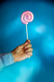 Colorful lollipop swirl on stick. Striped spiral multicolored pink candy on blue background, holding in hand sweet hard candy