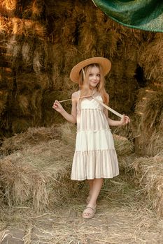 Romantic smiling tween girl in light sundress and straw hat standing on hayloft on background of haystack on sunny summer day. Country vacation concept