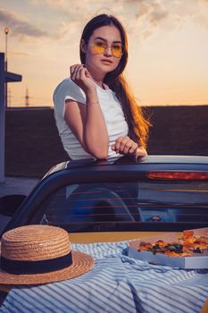 Young gorgeous woman in sunglasses, white t-shirt is posing in yellow car roadster with pizza, striped cloth and hat on its trunk. Fast food, unhealthy nutrition. Summer evening. Close up