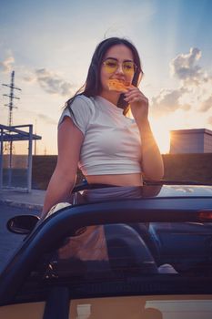 Young brunette female in sunglasses and white t-shirt is eating slice of pizza while posing standing in yellow car cabriolet. Fast food, unhealthy nutrition. Summer sunset. Close up