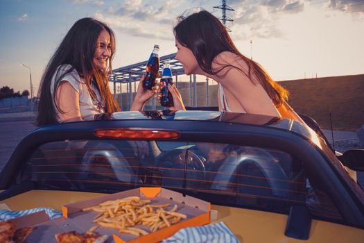 Happy women in casual outfit are smiling, cheering with soda in glass bottles, posing in yellow car with french fries and pizza on its trunk. Fast food. Summer evening. Close up, copy space, mock up